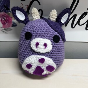 May include: A purple crocheted cow plush toy with white spots and black eyes. The cow has a purple and white crocheted nose and a purple crocheted patch on its head.