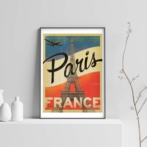 Op de afbeelding: Vintage reisposter met de Eiffeltoren in Parijs, Frankrijk. De poster heeft een rood-wit-blauwe kleurstelling en toont een vliegtuig dat overvliegt. De tekst "Paris France" is in zwart gedrukt aan de onderkant van de poster.