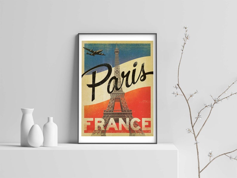 Op de afbeelding: Vintage reisposter met de Eiffeltoren in Parijs, Frankrijk. De poster heeft een rood-wit-blauwe kleurstelling en toont een vliegtuig dat overvliegt. De tekst "Paris France" is in zwart gedrukt aan de onderkant van de poster.
