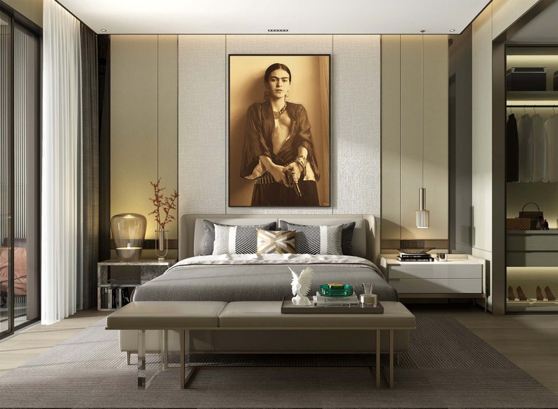 May include: A modern bedroom with a large bed, a bench at the foot of the bed, and a framed picture of Frida Kahlo on the wall. The bed has a gray headboard and a gray comforter. The bench is upholstered in a light brown fabric. The picture is in a black frame and features a sepia-toned image of Frida Kahlo holding a gun.