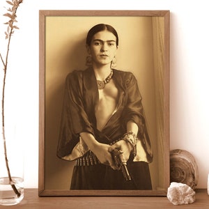 May include: A black and white photograph of a woman wearing a dark robe and holding a gun. She is wearing a necklace and bracelets. The photo is framed in a wooden frame.