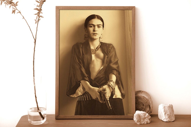May include: A black and white photograph of a woman wearing a dark robe and holding a gun. She is wearing a necklace and bracelets. The photo is framed in a wooden frame.