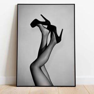 May include: Black and white framed print featuring a pair of legs in sheer black stockings and high-heeled shoes. The legs are positioned upwards, with the heels of the shoes pointing towards the top of the frame. The image is a study in contrasts and shadows.