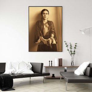 May include: A black and white portrait of Frida Kahlo holding a gun. She is wearing a dark robe and a necklace. The background is a light brown color.