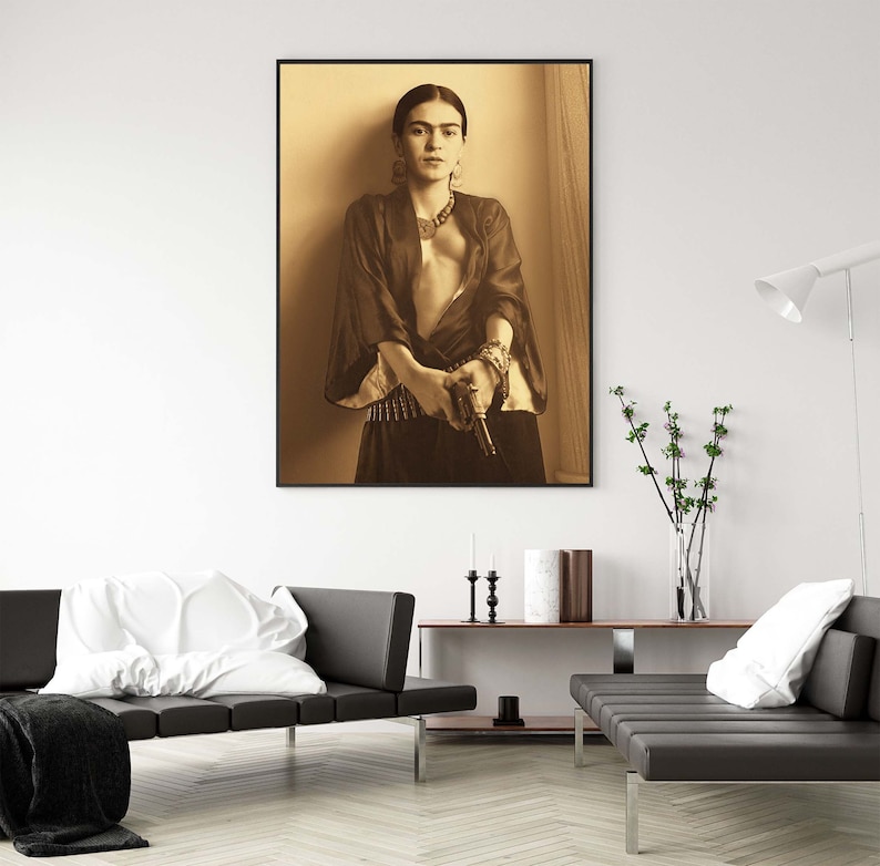 May include: A black and white portrait of Frida Kahlo holding a gun. She is wearing a dark robe and a necklace. The background is a light brown color.
