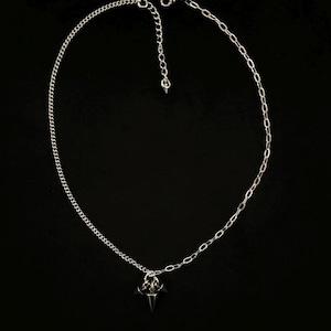 May include: A silver-colored necklace with a small, pointed pendant. The necklace has two chain styles: a curb chain and a cable chain. The pendant is a dark gray color and has a geometric design. The necklace is displayed against a black background.