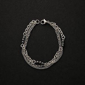 May include: Silver-toned bracelet featuring multiple chain strands and small, dark beads. The bracelet has a lobster clasp closure. The jewelry is displayed against a dark background, highlighting the metallic shine of the chains.
