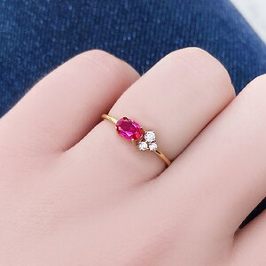 May include: A gold ring featuring a single oval-cut vibrant pink gemstone set horizontally next to a cluster of three small, round, clear white stones. The band is thin and polished gold, creating a delicate and refined appearance on the finger.