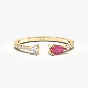May include: A gold-colored open ring with a white diamond and a red pear-shaped gemstone. The ring band is encrusted with small, clear gemstones. The ring is set against a white background.