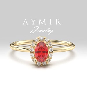 以下が含まれることがあります： 鮮やかな赤い楕円形の宝石をあしらったゴールドリング。小さな透明な宝石のハローで囲まれています。リングバンドは細く磨かれています。「AYMIR Jewelry」の文字が背景に表示されています。