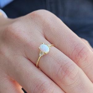Könnte beinhalten: Ein goldener Ring mit einem ovalen weißen Opal-Edelstein und kleineren weißen Edelsteinen auf der Ringschiene.