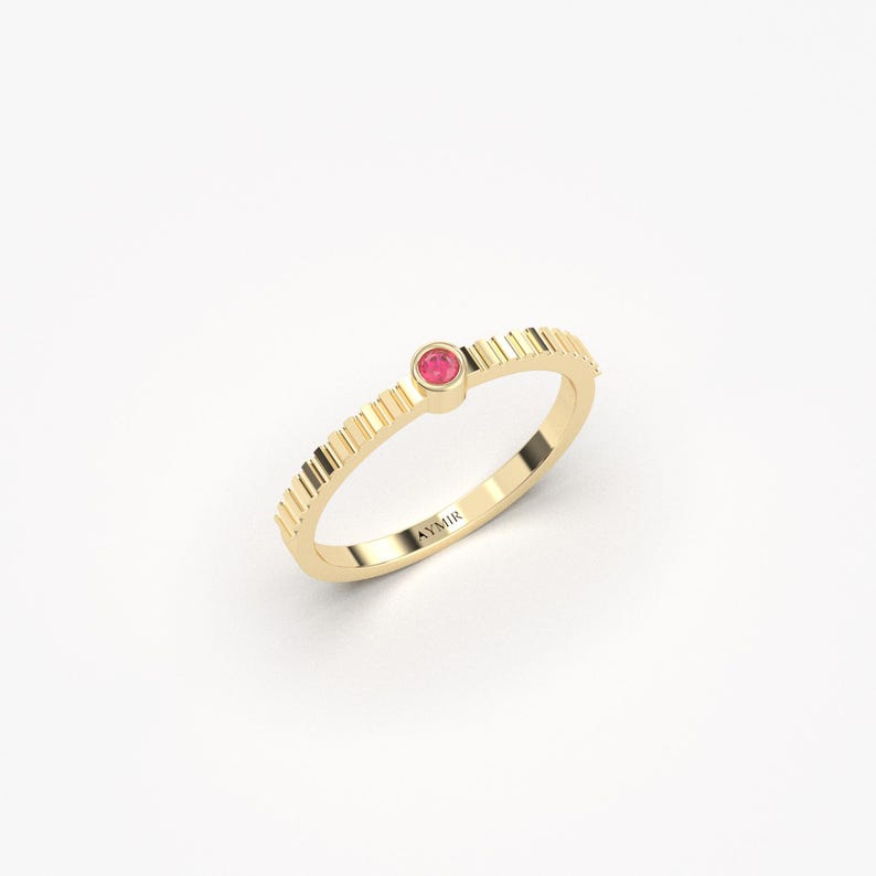 Pu&ograve; includere: Un anello d'oro con una piccola gemma rosa e rotonda. La fascia dell'anello presenta un design strutturato e scanalato. L'anello &egrave; posto su uno sfondo bianco. Il nome del marchio "AYMIR" &egrave; inciso sull'anello.