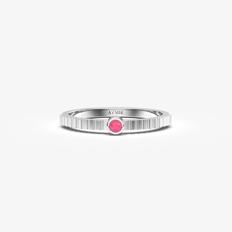 Pu&ograve; includere: Un anello d'argento con una pietra preziosa rosa al centro. La fascia ha un design a coste. La parola "AYMIR" &egrave; incisa sull'anello.