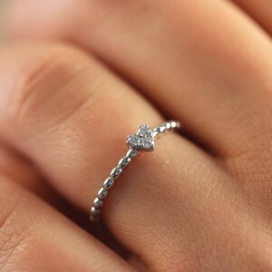 May include: A delicate silver ring with a small heart-shaped diamond accent. The ring is made of a beaded band.
