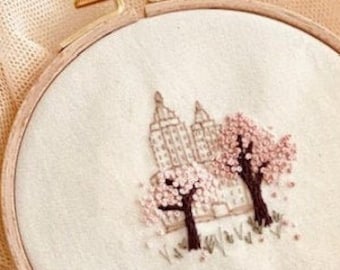 May include: A white embroidery hoop featuring a hand-stitched design of a city skyline with two pink cherry blossom trees. The embroidery is on a white fabric background.