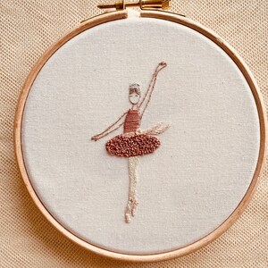 May include: Embroidered hoop art featuring a ballerina in a brown dress with a white bodice. The ballerina is in a dancing pose with her arms raised above her head.