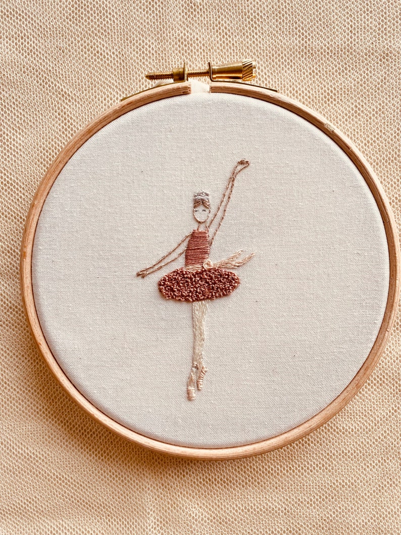 May include: Embroidered hoop art featuring a ballerina in a brown dress with a white bodice. The ballerina is in a dancing pose with her arms raised above her head.