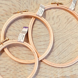May include: Three wooden embroidery hoops of varying sizes. The hoops are made of light-colored wood and feature brass-colored tightening screws. Each hoop has a label attached. The hoops are arranged on a textured, tan surface.
