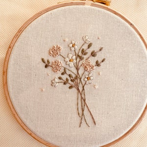 May include: A close-up of a hand-stitched embroidery hoop with a floral design. The embroidery features white and cream flowers with green leaves on a beige fabric background.