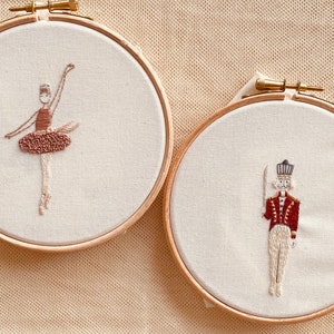 May include: Two embroidery hoops with hand-stitched designs. The left hoop features a ballerina in a brown tutu, while the right hoop depicts a toy soldier in a red uniform.