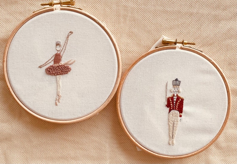 May include: Two embroidery hoops with hand-stitched designs. The left hoop features a ballerina in a brown tutu, while the right hoop depicts a toy soldier in a red uniform.