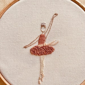 May include: Embroidered ballerina in a brown dress with a white headband, on a white fabric background in a wooden embroidery hoop.