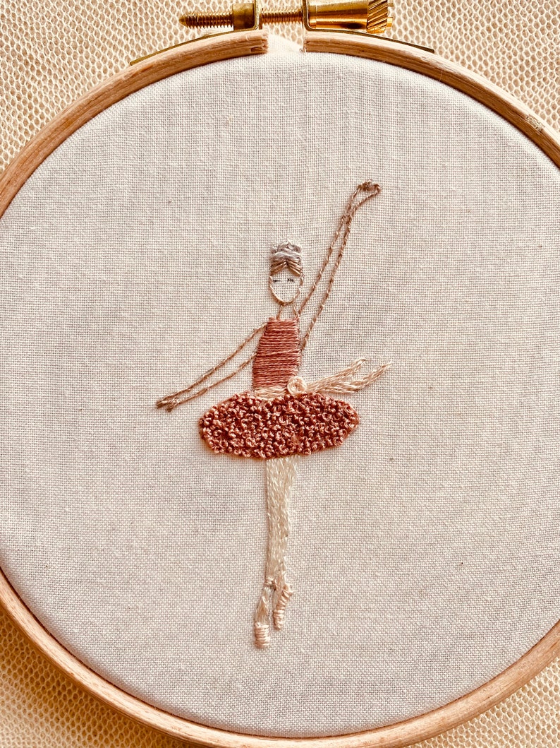 May include: Embroidered ballerina in a brown dress with a white headband, on a white fabric background in a wooden embroidery hoop.