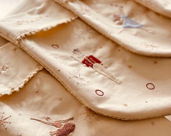Puede incluir: Calcetines navideños color crema con bailarinas de ballet y copos de nieve bordados. Cada calcetín presenta un borde festoneado y bordados detallados en tonos rojos, marrones y azules. Una decoración festiva.
