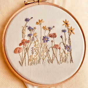 May include: A close-up of a circular embroidery hoop with a beige fabric background. The fabric is embroidered with a variety of colourful flowers, including purple, yellow, and orange.