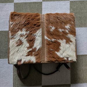 Puede incluir: Un bolso de cuero de vaca marrón y blanco con una correa larga. El bolso está cerrado y tiene una solapa con una hebilla.