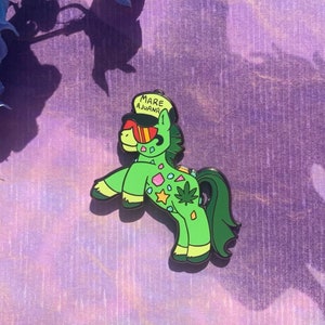 May include: Green cartoon unicorn pin with a marijuana leaf design and the words "Mare Ajwana" on its hat.