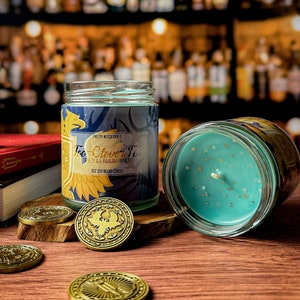 Puede incluir: Un tarro de vela azul y dorado con el texto "Too Clever To" y "Tres Slas Ravkan Loyalty" en la etiqueta. La vela está rodeada de monedas de oro con un diseño de pájaro.