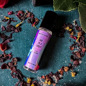 May include: A small, clear glass bottle with a black rollerball top. The bottle is filled with a pink and purple liquid. The label on the bottle reads "Pretty Witch Craft Give Me A Thrust Lust Desire 10ml Perfume Oil". The bottle is surrounded by red rose petals and green leaves on a teal background.