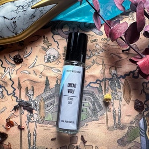 May include: A small glass bottle with a black roller ball top. The bottle is labeled "Dread Wolf Tears & Lies" and "10ml Perfume Oil" in white text on a blue background. The bottle is sitting on a brown and white patterned surface with a skeleton and other decorative elements.