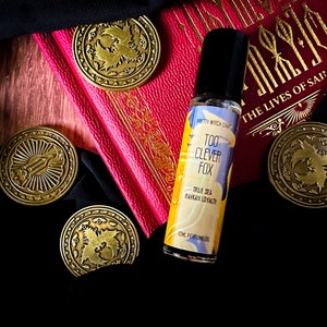 Puede incluir: Un pequeño frasco de vidrio de aceite de perfume con una tapa negra. La etiqueta es azul y amarilla con el texto "Too Clever Fox" y "True Sea Ravkan Loyalty". El frasco está rodeado por cuatro fichas redondas de metal dorado con un diseño de un pájaro.