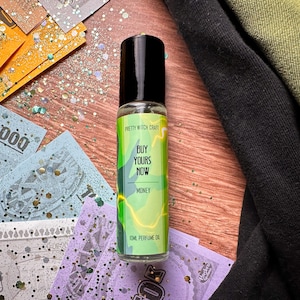 May include: A clear glass bottle with a black cap and a green and yellow label. The label reads "Pretty Witch Craft", "Buy Yours Now", "Money", and "10ml Perfume Oil".