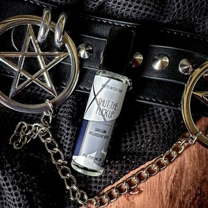 以下が含まれることがあります： 「Pretty Witch Craft」と「Shut the Fuck Up」と書かれた白黒のラベルが付いた、小さなガラス製の香水オイルボトル。ラベルの下には「Sarcasm Hellhound Bite」と「Roll Perfume Oil」の文字が書かれている。ボトルは、銀色のスタッズと銀色のチェーンが付いた黒い革のベルトの上に置かれている。