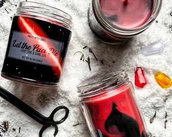 May include: Two red soy blend candles with black and white galaxy designs. The candles are in glass jars with the text "Let the Past Die" and "Light Side & Dark Side" on the front. The candles are sitting on a white surface.
