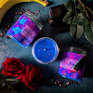 May include: Two blue glass candle jars with pink and purple labels that read "Give Mea Trust" and "Give Mea Trust Bourbon & Cologne" on a dark blue background with red roses, a banana, and a dark brown bottle.