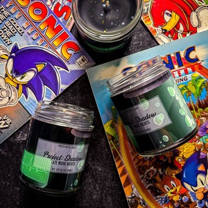 May include: Three black soy blend candles with labels that read "Project: Shadow Late Work Nights" and "Black & Shadow Late Work Nights". The candles are sitting on top of comic books featuring the video game character Sonic the Hedgehog.