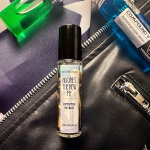 Puede incluir: Un pequeño frasco de vidrio de aceite de perfume con una tapa negra. La etiqueta es blanca con texto negro que dice "Pretty Witch Craft", "No One's The New Me", "Patriotism Payback" y "10ml Perfume Oil".