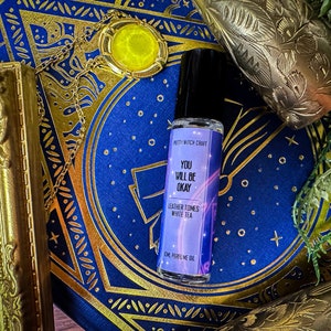 May include: A perfume oil bottle with a black cap and a label that reads "YOU WILL BE OKAY." The label also lists "Leather Tones White Tea." The bottle is set on a blue surface with gold accents and a gold-framed object.