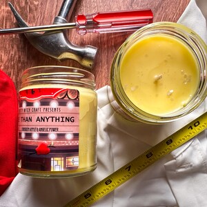 May include: A yellow candle in a glass jar with a label that reads "Witch Craft Presents More Than Anything Forbidden Apple & Angelic Power".
