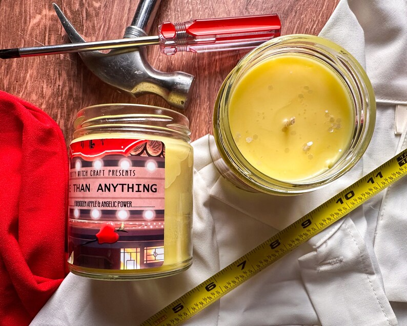 May include: A yellow candle in a glass jar with a label that reads "Witch Craft Presents More Than Anything Forbidden Apple & Angelic Power".