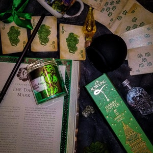 May include: A green and gold candle with a dark green snake design. The candle is in a glass jar with a black lid. The candle is sitting on a book with the text "Chapter Nine: The Dark Mark" visible. The book is open and has a green and gold design on the cover. There is a green and gold box with the text "J.K. Rowling Harry Potter and the Chamber of Secrets" visible. The box is sitting on a black surface. There are also some playing cards and other objects in the image.
