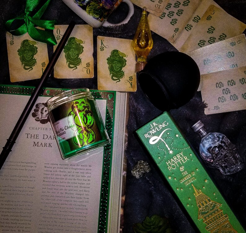 May include: A green and gold candle with a dark green snake design. The candle is in a glass jar with a black lid. The candle is sitting on a book with the text "Chapter Nine: The Dark Mark" visible. The book is open and has a green and gold design on the cover. There is a green and gold box with the text "J.K. Rowling Harry Potter and the Chamber of Secrets" visible. The box is sitting on a black surface. There are also some playing cards and other objects in the image.
