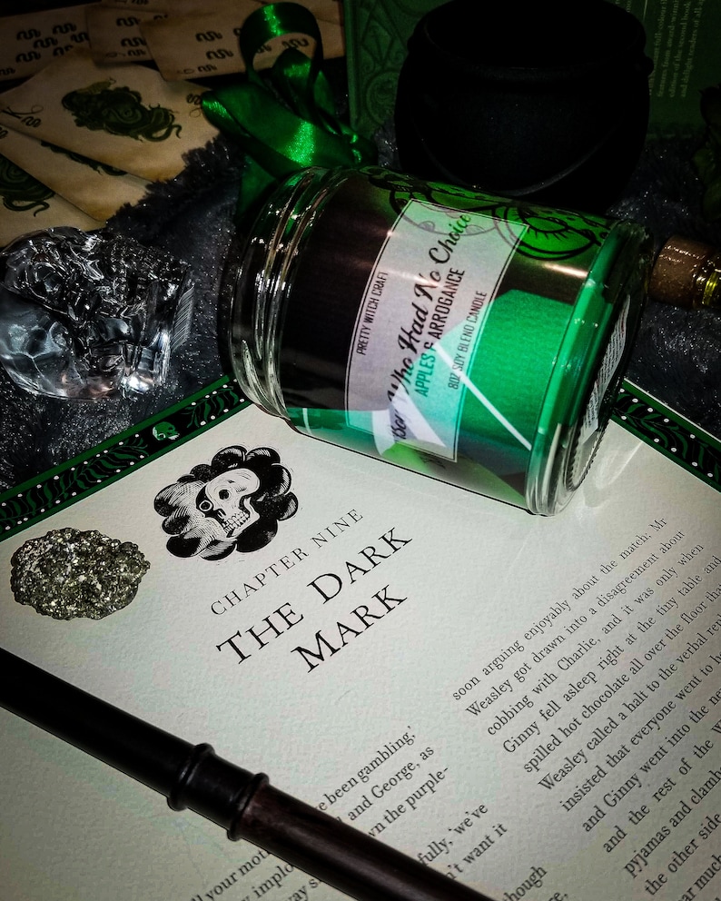 May include: A green candle with the text "Sor Tho Had No Choice Apples & Arrogance 8oz Soy Blend Candle" on the label. The candle is sitting on a book with the title "The Dark Mark" and a black wand.