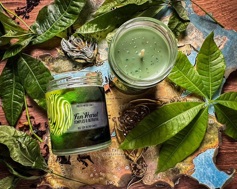 May include: A green soy blend candle in a glass jar with a label that reads "Fen'Harel Temples & Betrayal". The candle is surrounded by green leaves and a map.