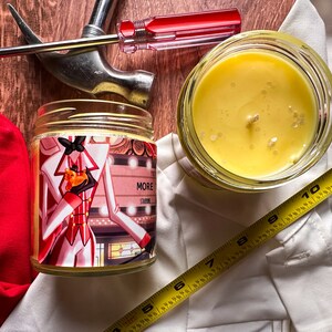 May include: Two glass candle jars containing yellow wax. One jar features a label with a cartoon illustration of a person in a red and white suit holding a small orange cat. The label also includes the text "More Shine".