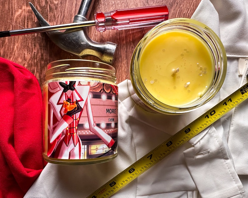 May include: Two glass candle jars containing yellow wax. One jar features a label with a cartoon illustration of a person in a red and white suit holding a small orange cat. The label also includes the text "More Shine".